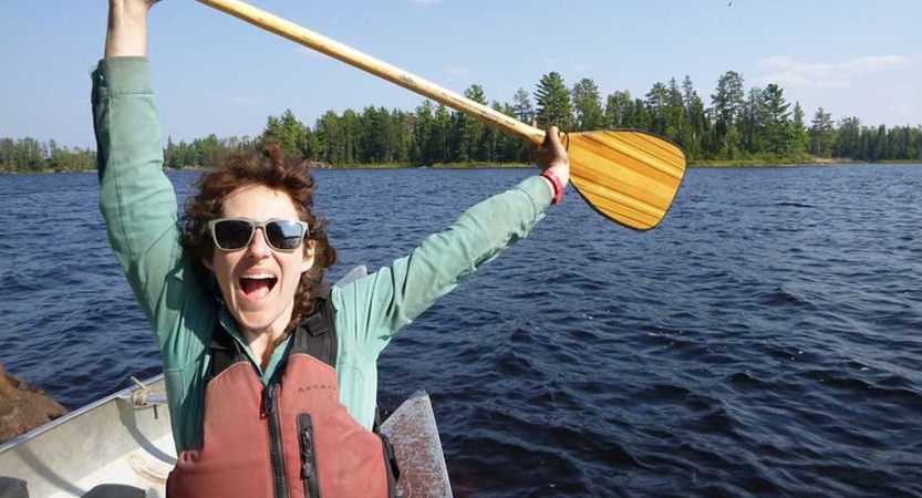 A person wearing a life jacket sits in a canoe and raises their paddle above their head in celebration. 